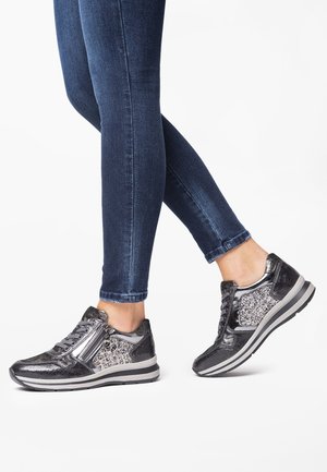 Stopy w metalicznych, ciemnoszarych sneakersach z błyszczącymi panelami i bocznymi zamkami, w połączeniu z obcisłymi, ciemnoniebieskimi dżinsami na białym tle.