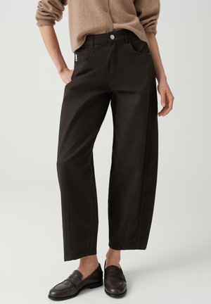 Persona che indossa pantaloni neri a gamba larga, maglione marrone con bottoni e mocassini in pelle marrone scuro, in piedi con una mano nella tasca.
