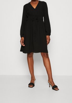 Robe noire à manches longues, décolleté en V et ceinture élastique à la taille. Tissu lisse, longueur au genou. Portée avec des sandales à talons noires.
