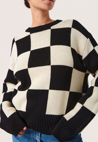 Schwarzer und cremiger karierter Pullover mit geripptem Kragen und Bündchen, der eine lockere Passform und Stricktextur aufweist.