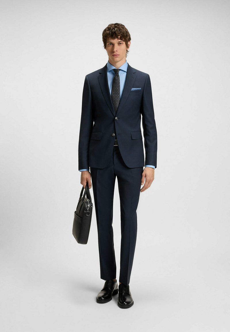 Marineblauer Anzug mit tailliertem Blazer und schmal zulaufenden Hosen, kombiniert mit einem hellblauen Hemd und einer gemusterten Krawatte; hält eine schwarze Tasche.