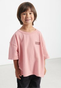 Lyserød oversized bomulds t-shirt med korte ærmer, der har et frontlogo "AMERICAN VINTAGE". Båret med mørke fløjlsbukser.