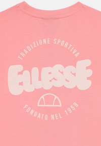 Rožinė medvilninė marškinėlė su didele balta grafika, kurioje yra užrašai "Ellesse" ir "Tradizione Sportiva" apvaliais šriftais.