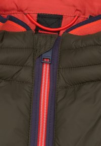 Donkergroen pufferjack met een olijfgroen buitenkant, rode voering en een contrasterende felrode rits op een donkerpaarse flap.