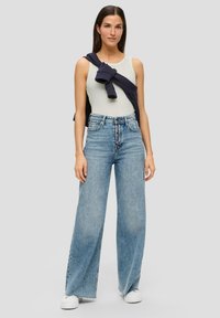 Canotta verde chiaro, jeans in denim blu chiaro a gamba larga e vita alta, con cinque bottoni frontali, indossata con un maglione blu navy drappeggiato sulle spalle. Sneakers bianche.