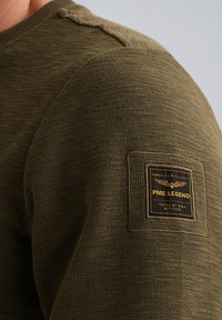 Suéter de punto en verde oliva con un diseño texturizado y acanalado. Parche cuadrado en el hombro con detalles de marca en negro y dorado.