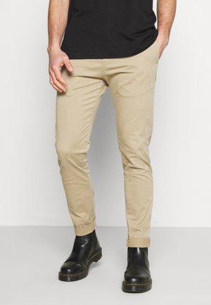 Mann trägt beige, schmal geschnittene Hose, an den Bündchen hochgekrempelt, und schwarze Lederstiefeletten, steht mit einer Hand in der Tasche.