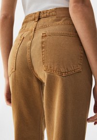 Pantalon en denim marron taille haute avec une coupe fuselée. Il présente deux poches arrière et un tissu texturé avec des détails de couture visibles.