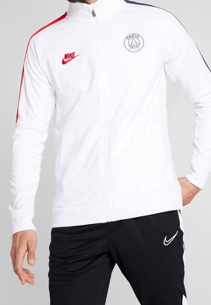 Muž nosící bílou bundu Paris Saint-Germain od Nike s červenými a modrými pruhy na rukávech a černé kalhoty Nike s bílým logem Swoosh.