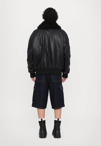 Zwarte leren bomberjack met een textuurafwerking, uitgerust met een zwarte nepbont kraag. Gecombineerd met donkere denim cargo shorts en zwarte laarzen.