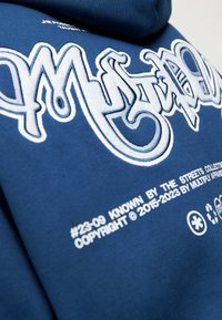 Felpa con cappuccio blu con grande scritta bianca ricamata. Include una piccola etichetta stampata con simboli di copyright e riciclaggio. Tessuto dalla trama liscia.