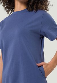 Femme aux cheveux bouclés portant une chemise simple à manches courtes bleue, une main placée dans une poche latérale sur un fond clair.
