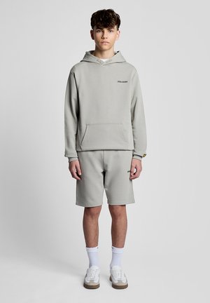 Adolescente in piedi, indossa una felpa con cappuccio grigio chiaro e pantaloncini abbinati con calzini bianchi e scarpe da ginnastica, sfondo neutro.