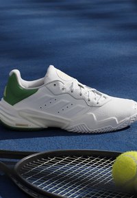 Scarpa da tennis bianca e verde su campo blu accanto a una racchetta da tennis nera e una pallina da tennis gialla.