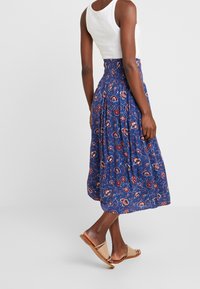 Blaues Blumen-Midi-Rock mit gesmoktem Taillenbund, der ein Muster aus roten und weißen Blumen auf einem strukturierten Stoff zeigt. Kombiniert mit beigen Sandalen.