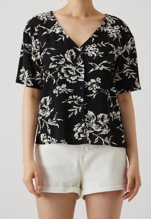 Personne portant un haut noir à manches courtes boutonné avec imprimé floral blanc et un short blanc retroussé, debout devant un fond uni.