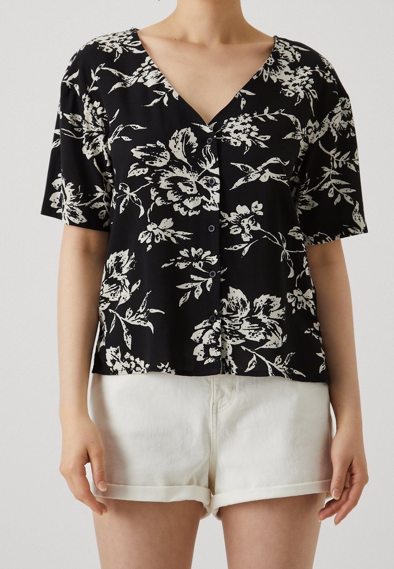 Personne portant un haut noir à manches courtes boutonné avec imprimé floral blanc et un short blanc retroussé, debout devant un fond uni.