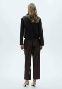 Veste courte en cuir marron avec manches longues, associée à un pantalon marron ample. Le mannequin porte des chaussures à talons jaunes. Design minimaliste, texture lisse.