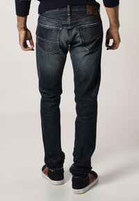 Tmavě modré džíny z denimu, fitted střih, pět kapes, vybledlá textura s kontrastním prošíváním a kožená nášivka na pásku.