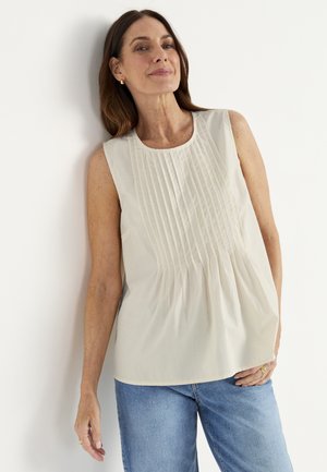 Kvinde, der læner sig op ad en hvid væg iført en ærmeløs cremefarvet bluse med folder foran og blå jeans, let smilende.