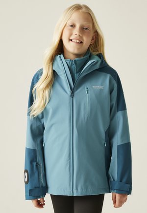 Jeune personne aux longs cheveux blonds portant une veste imperméable à fermeture éclair bleu clair et bleu marine avec une capuche.