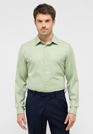 Camicia a maniche lunghe verde chiaro con colletto, davanti a un solo petto e colletto interno con motivo a contrasto. Indossata con pantaloni blu navy.