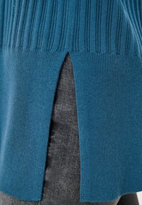Teal stickad tröja med ribbad textur, har sidoflikar, kombinerad med grå denim. Närbilden framhäver tyget och designen.