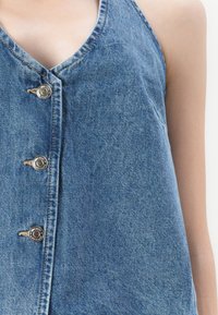 Denim brezrokavni top v svetlo modri barvi z V-izrezom, opremljen s srebrnimi pritisnimi gumbi in vidnimi šivi. Mehka tekstura, sproščen dizajn.