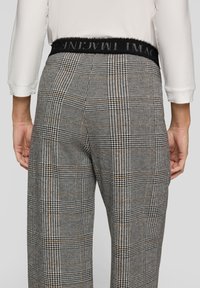 Person, die graue karierte, hochtaillierte Hosen mit einem schwarzen Bund und ein weißes langärmliges Oberteil trägt, von hinten gezeigt.