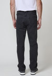 Hombre vestido con tejanos negros y zapatillas negras con suelas blancas, de pie de espaldas contra un fondo claro y liso.