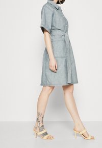 Kvinna som promenerar i en kortärmad jeansklänning med skärp och sidofickor, bär beige klackskor och har flera tatueringar på vänster fotled och fot.