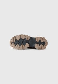 Suela de zapatilla deportiva con robusto caucho negro que presenta surcos profundos y secciones en marrón claro, diseñada para ofrecer tracción y durabilidad.