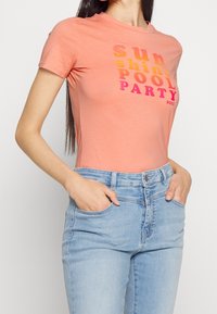 Peachfärgad t-shirt med kort ärm och djärv text "SUNSHINE POOL PARTY" i en gradient av pink och orange. Mjuk, figurnära bomullsmaterial.
