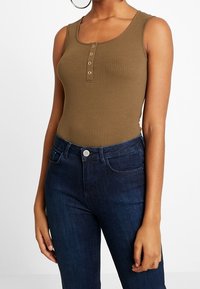 Olivgrön ribbad ärmlös bodysuit med rundad halsringning och tre metallknappar framtill, matchad med mörkblå jeans.