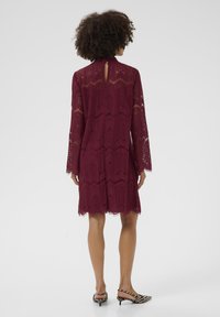 Vestido de encaje burdeos con mangas largas, dobladillo con puntilla y detalle en la espalda con abertura. Llevado con mules de tacón a rayas blancas y negras.