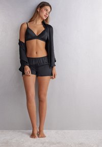 Svart satäng bralette och shorts-set. Braletten har v-ringning och justerbara band. Modellen bär en öppen svart skjorta och står på en ljus matta.