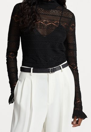 Femme portant un haut à manches longues en dentelle noire avec des motifs complexes, rentré dans un pantalon blanc plissé taille haute avec une ceinture noire.