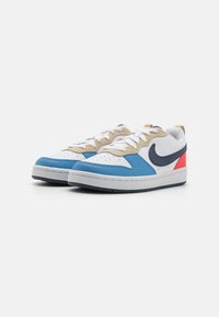 Nike sneakers featuring a white leather upper, beige accents, a blue side panel, and red detailing on the heel. Textured rubber sole.