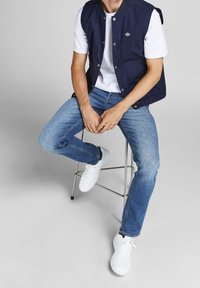 Navy blue sleeveless jacket over a white shirt, accompanied by light blue jeans and white sneakers, seated on a metal stool.