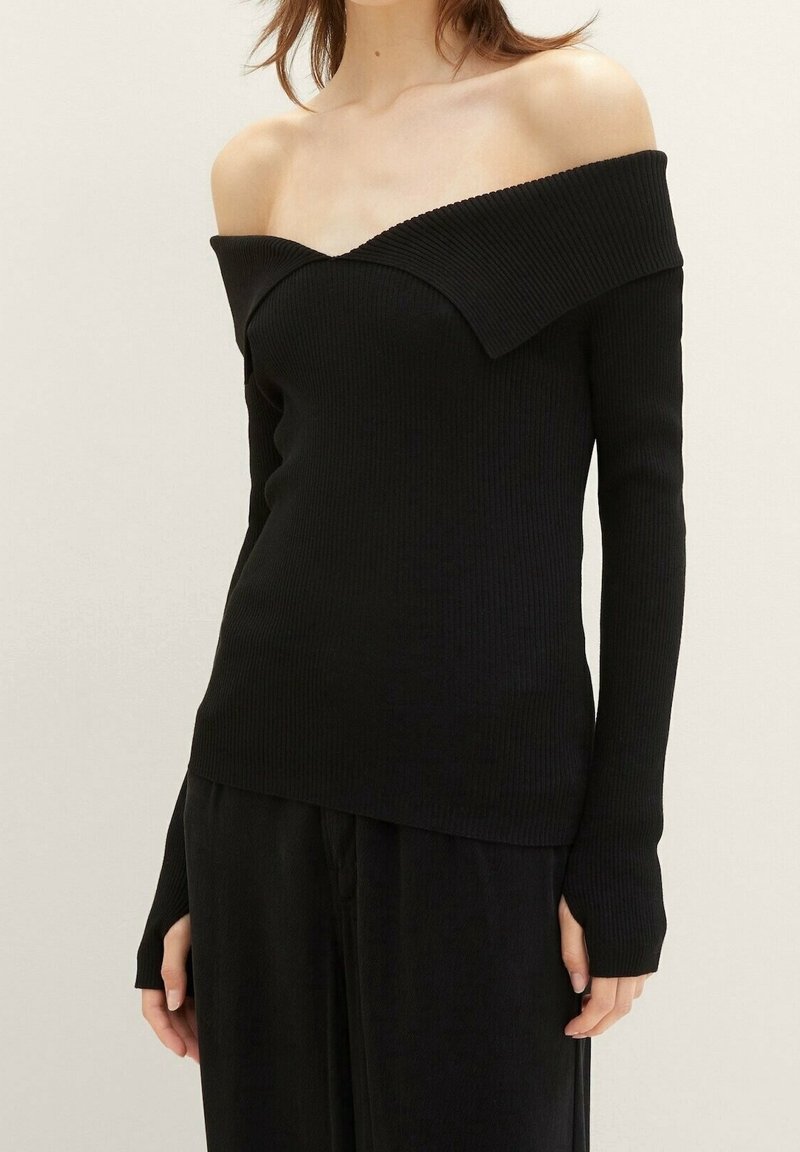 Femme portant un haut noir côtelé à manches longues, à épaules dénudées, associé à un pantalon noir ample sur un fond uni.