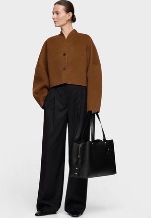 Mujer con chaqueta marrón grande abotonada, pantalones negros de pierna ancha, sosteniendo un gran bolso tote negro, posando frente a un fondo liso.