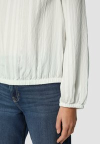 Witte, geribde lange mouw shirt met verzamelde manchetten en zoom, gecombineerd met donkerblauwe jeans. Materiaal lijkt lichtgewicht en gestructureerd.