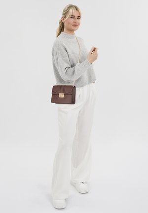 Maglione in maglia grigia con texture, pantaloni bianchi a gamba larga e borsa a tracolla marrone con dettagli dorati. La modella posa con un braccio alzato, indossando sneaker bianche.