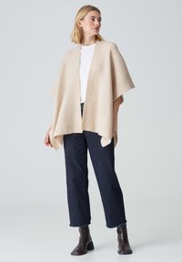 Poncho beige tricoté avec une texture côtelée, à l'avant ouvert et aux manches amples, porté sur une chemise blanche et un jean en denim foncé. Des bottes à chevilles marron complètent le look.