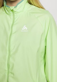 Chaqueta verde claro con cremallera frontal, cuello alto y un interior azul en contraste. Presenta el logo de Odlo en blanco en el pecho. Textura suave.