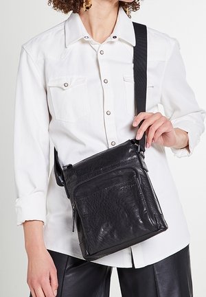Femme portant une chemise blanche à boutons et un pantalon en cuir noir, tenant un sac bandoulière en cuir noir avec une poche avant zippée.