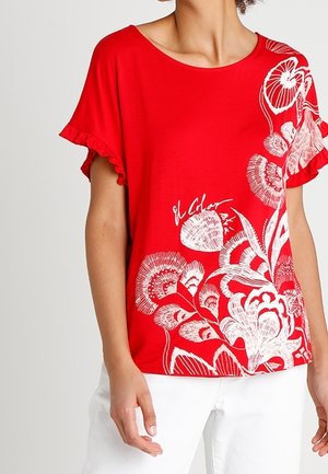 Kvinde iført en rød top med korte ærmer med hvidt blomsterprint og flæseærmer, kombineret med hvide bukser mod en ensfarvet baggrund.