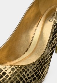 Chaussure à talon haut dorée avec un motif texturé effet peau de serpent. Intérieur avec une finition métallique lisse. Forme structurée avec un bout pointu.