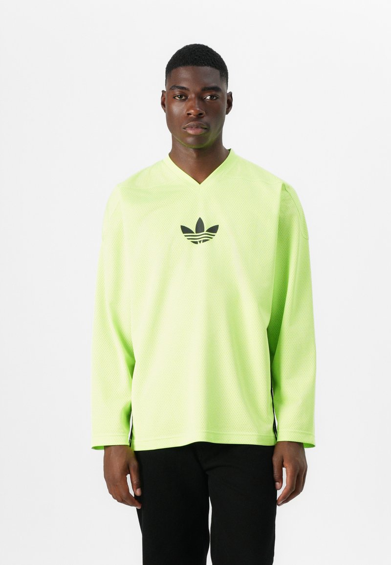 Neon zelena dolgosleeve majica iz mrežaste tkanine z V-izrezom in črnim logotipom na prsih. Ima stranske razpoke na robu.