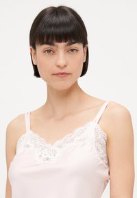 Rózsaszín selyem camisole, csipkés díszítéssel a nyakkivágás mentén és vékony pántokkal, sima textúrával és V-alakú kialakítással.
